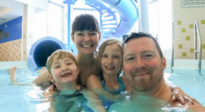 A selfie of a family of four smiles and embraces each other in an indoor pool. A large blue waterslide in the background.