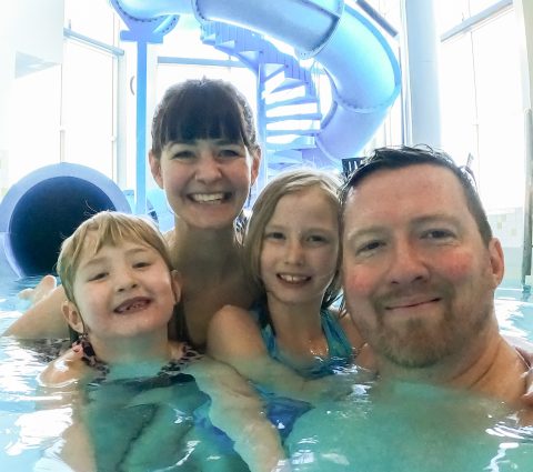 Selfie d'une famille de quatre personnes souriantes et enlacées dans une piscine intérieure. Un grand toboggan bleu en arrière-plan.