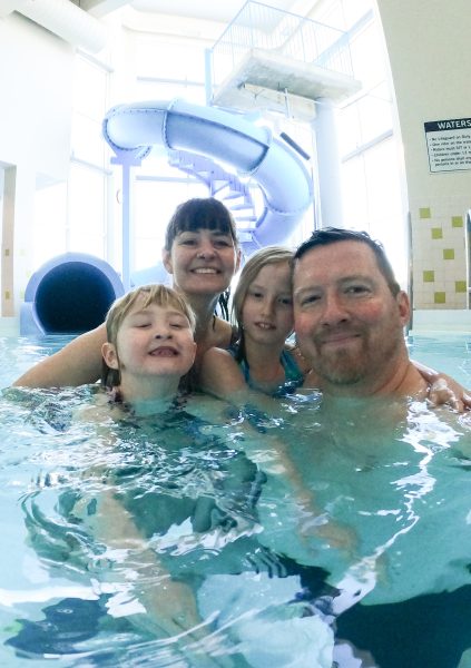 Selfie of the family of four in the pool at the hotel. A large blue waterslide is visible in the background.