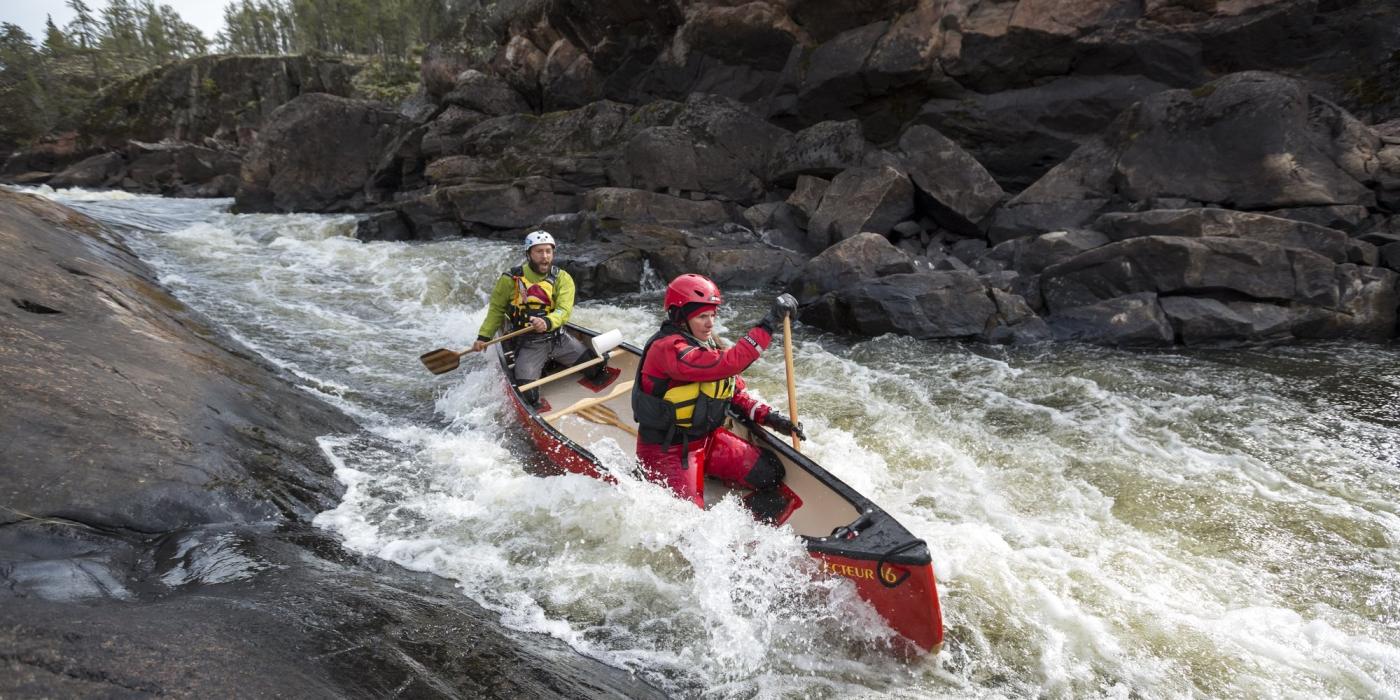 Deux personnes parcourant des rapides dans un canoë rouge. Il y a de hautes parois rocheuses de part et d'autre.