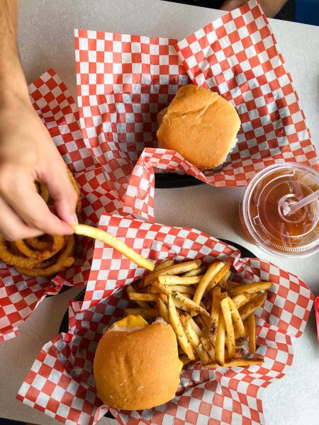 A hand reaches over onion rings, burgers on a table to pick up a french fry.