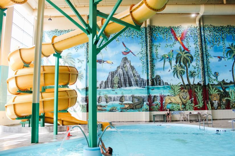 Isang dilaw, twisty waterslide at pool area sa backdrop ng isang mural na may mga tropikal na halaman at kakaibang hayop