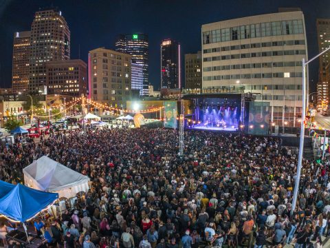 Nighttime block party with a massive crowd gathered around an outdoor stage amid city lights and surrounding tall buildings.