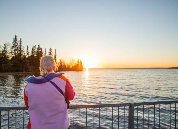 Eine Frau steht an einem Geländer und beobachtet den Sonnenuntergang über einem See.