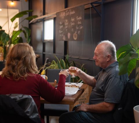 Deux personnes dégustent un repas et trinquent avec des boissons à l'intérieur d'une salle de dégustation de cidrerie accueillante et remplie de plantes.