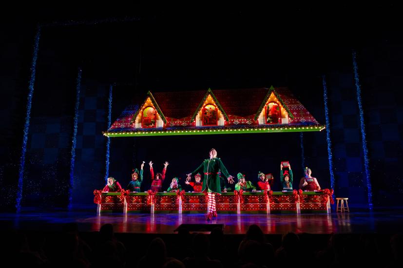 Buddy le lutin se tient au centre de la scène et ses compagnons lutins chantent derrière lui. Rideau de fond bleu foncé avec des lumières rouges et vertes.