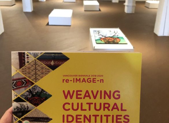 Una mano sostiene una tarjeta en la que se lee "Tejiendo identidades culturales", en primer plano, y detrás, unos tejidos colocados cerca del suelo.