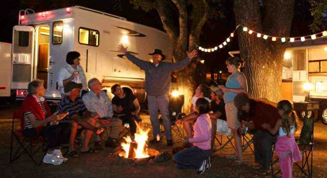 Una familia numerosa se reúne alrededor de una hoguera nocturna con sus caravanas al fondo.