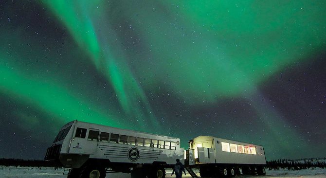 Une personne debout dans la toundra devant les véhicules du restaurant Dan's Diner sous les aurores boréales.