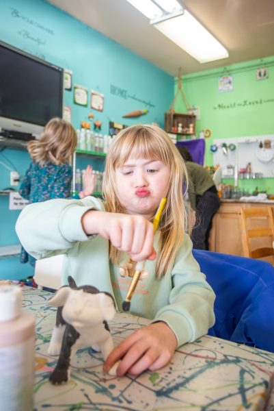 One little girl is concentrating while painting black onto a dog figure, her sister runs by in the background.