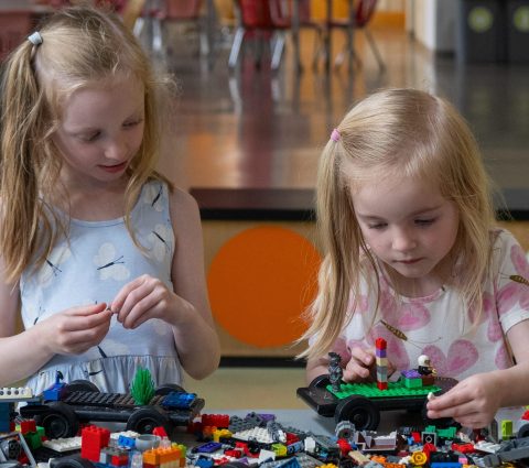 Two young children play with colorful building blocks on a table in a bright, spacious indoor play area.