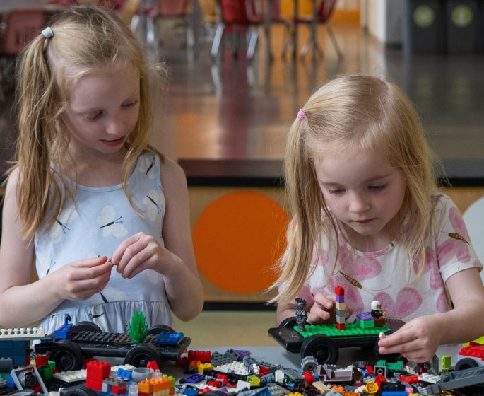 Two young children play with colorful building blocks on a table in a bright, spacious indoor play area.