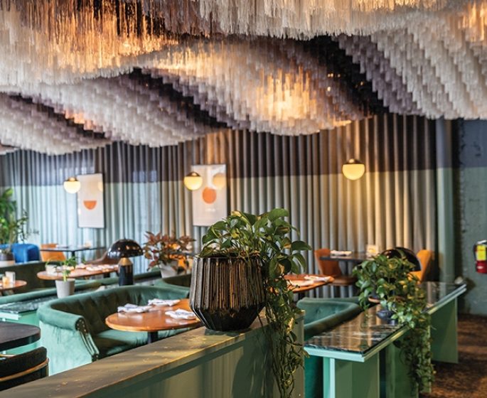 Intérieur de restaurant élégant, avec des sièges verts, des plantes décoratives et un plafond au design unique orné d'éléments suspendus.