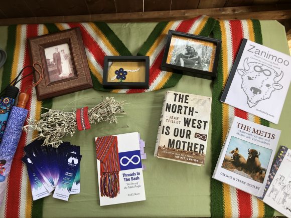 Eine Sammlung von Gegenständen auf einem Tisch - gerahmte Schwarzweißfotos, eine gerahmte Perlenblume, eine Metis-Schärpe und fünf Bücher