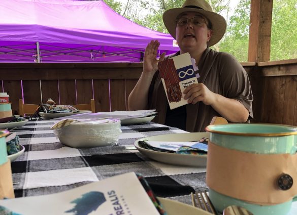 Eine Frau sitzt an einem Tisch, mit leicht geöffnetem Mund und erhobener Hand, mitten im Satz. In der Hand hält sie ein Buch mit dem Titel Threads in the Sash
