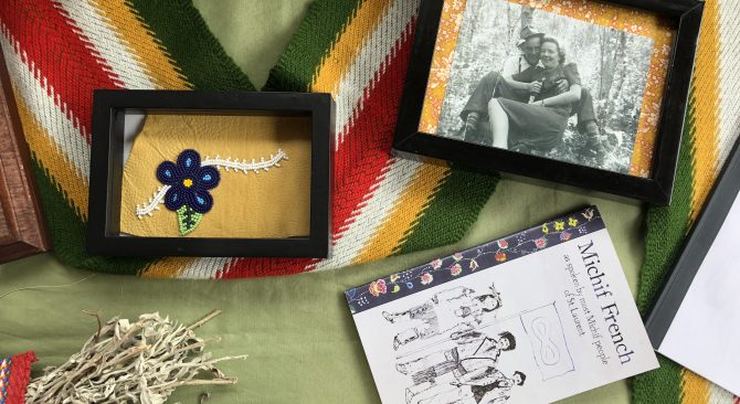 Una mesa con varios objetos: una foto en blanco y negro enmarcada, una flor de abalorios enmarcada, una faja Metis y un libro.