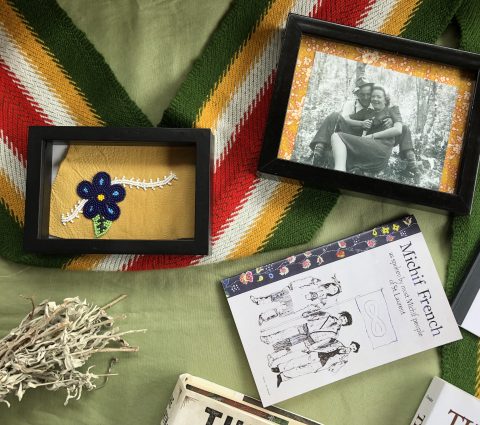Una mesa con varios objetos: una foto en blanco y negro enmarcada, una flor de abalorios enmarcada, una faja Metis y un libro.