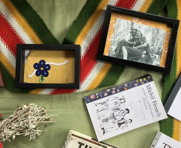 A table with various objects on it - a framed black and white photo, a framed beaded flower, a Metis sash and a book