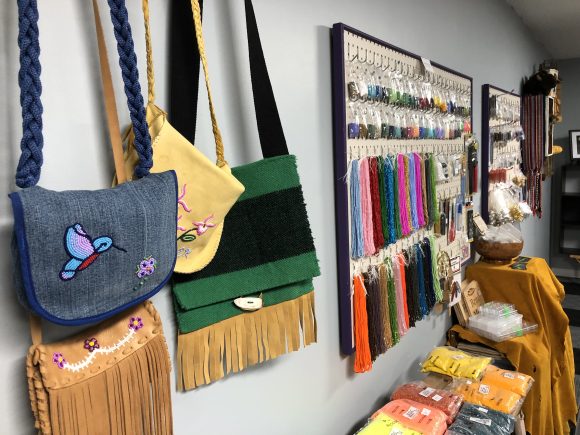 Vier Taschen mit Perlenkunst hängen an einer Wand. Weiter unten an der Wand hängen Schnüre mit vielen bunten Perlen