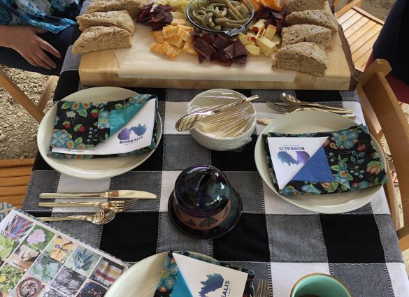 Ein mit Tellern und Stoffservietten gedeckter Tisch mit einem großen Holzbrett mit Bannock, Käse, Dörrfleisch und Essiggurken