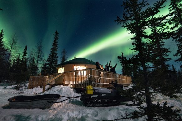 People standing on a deck outside a yurt at night, watching the northern lights dance overhead at Nanuk Lodge.