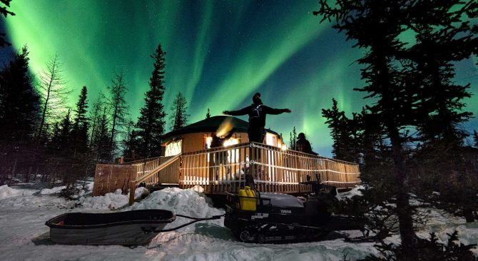Drei Personen auf der Terrasse einer Jurte, die das tanzende Nordlicht in der Nähe von Churchill beobachten.