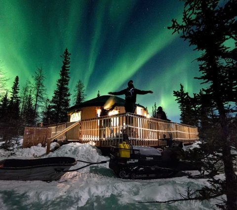 Three people on a deck at a yurt, watching the northern lights dance overhead near Churchill.