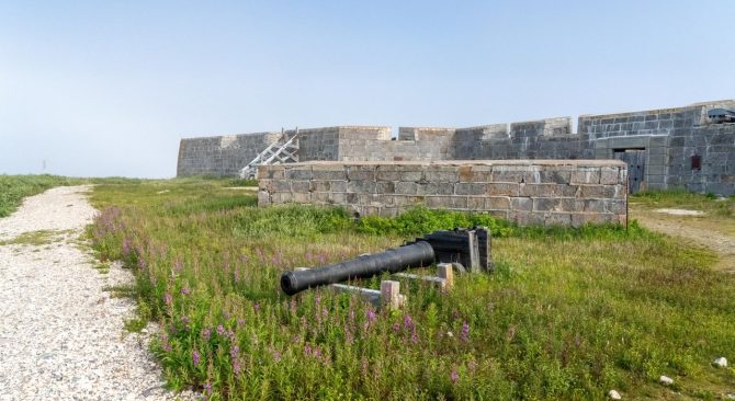 A view at a canon in the grass, a gravel path and the walls of Prince of Wales Fort in Churchill, Manitoba, against blue sky