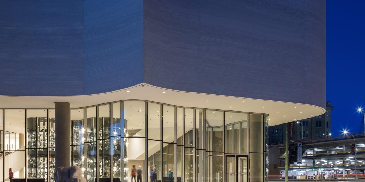 Exterior of Qaumajuq at night, large glass windows at base of building and wave-like cement facade on top