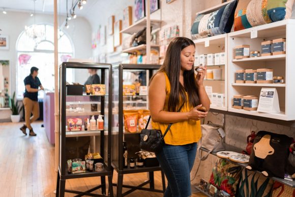 Una persona con camisa amarilla huele una pequeña vela frente a hileras de productos locales en Tara Davis Studio Boutique.