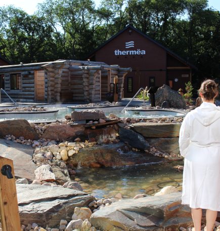 A lady in a white robe standing overlooking the outdoor pools and spa at Thermëa by Nordik Spa-Nature