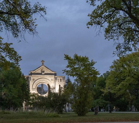 St. Boniface Cathedral illuminated at dusk during late summer. The building is bordered by green foliage.