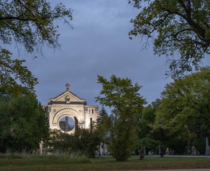La cathédrale Saint-Boniface illuminée à la tombée de la nuit, à la fin de l'été. Le bâtiment est entouré d'une végétation verdoyante.