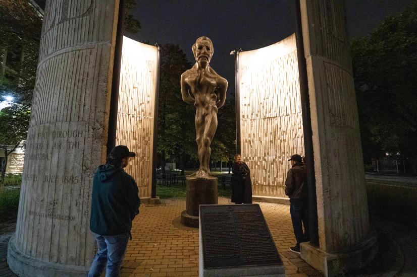 La statue en bronze de saint Boniface et de Louis Riel, illuminée la nuit, avec trois personnes qui l'observent.