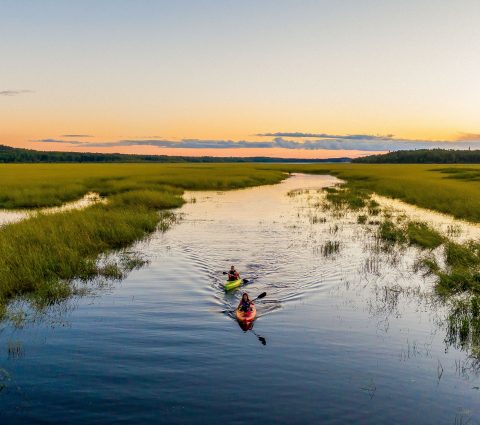 Dalawang kayaker na nag-e-enjoy sa sunset paddle sa isang malinaw na kalmadong gabi.