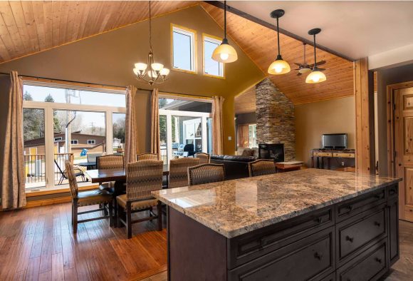 Nakatingin sa isang kitchen island papunta sa open dining area na may vaulted ceiling at floor to ceiling na mga bintana.