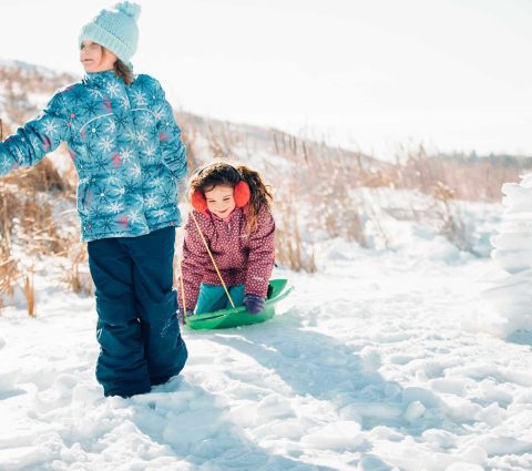 Two kids pulling each other on the snow in a green toboggan.