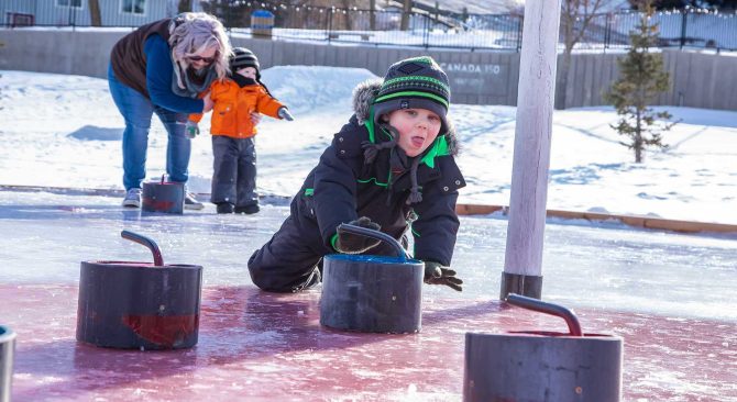 A young child on the ice curling, an adult and child play in the background.