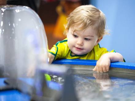 Young child leaning over a water activity table, smiling and reaching into the water while exploring a hands-on exhibit.