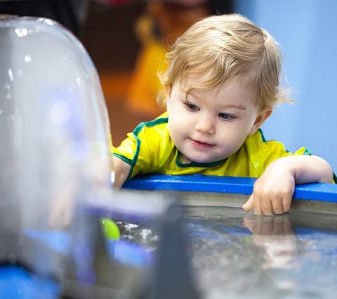 Young child leaning over a water activity table, smiling and reaching into the water while exploring a hands-on exhibit.