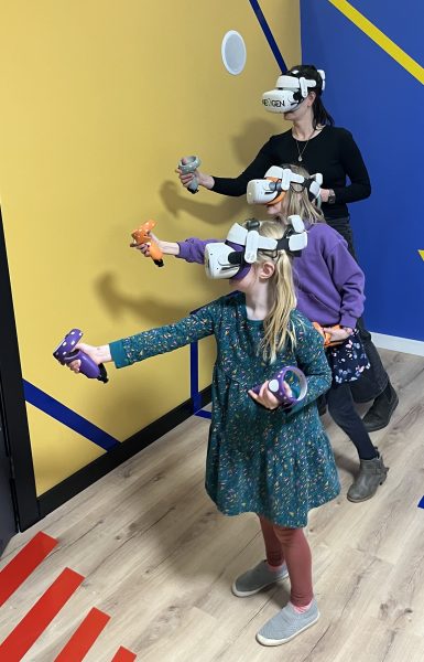 Two little girls and their mother stand in a row each with their VR goggles and blasters pointed out.
