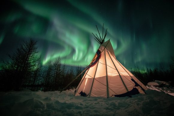 A tipi beneath the northern lights, lit from within.