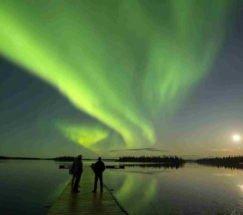 Dos personas de pie en un muelle durante la luna llena, observando la aurora boreal.