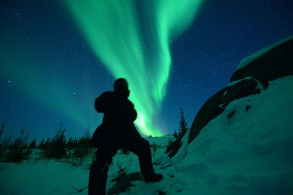 A person standing next to a rocky ridge looking up as the green northern lights dance overhead.