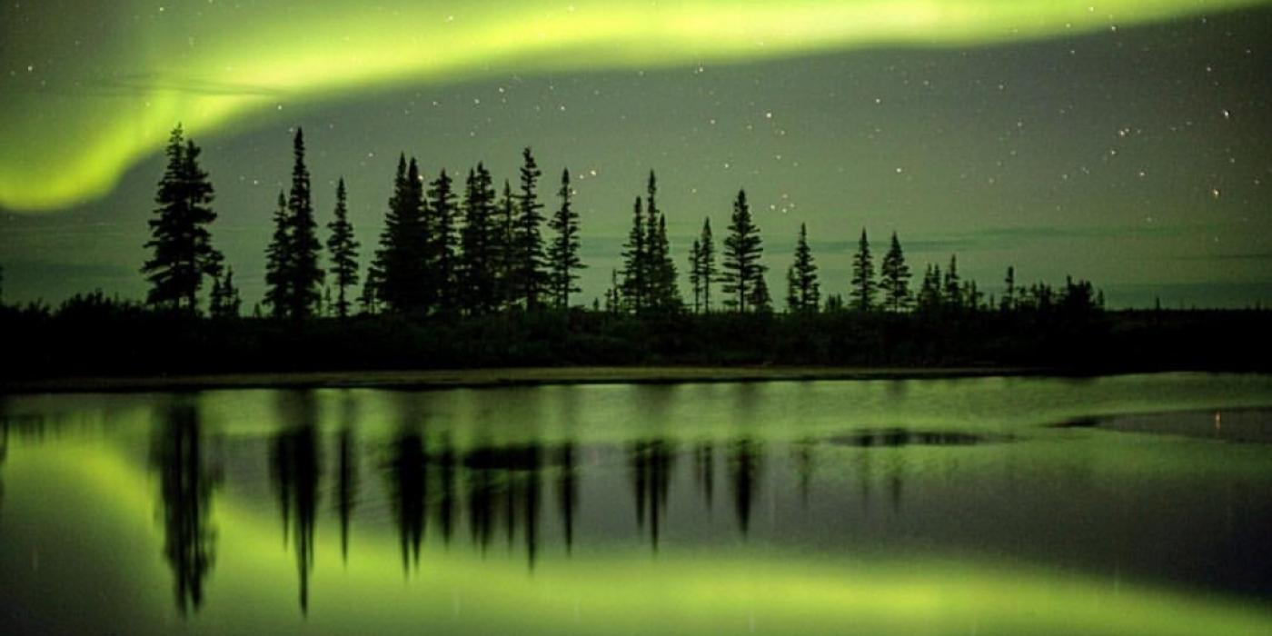 Northern lights fill the sky, trees reflect of the glass like ice. Where nature's dance illuminates the Arctic skies.