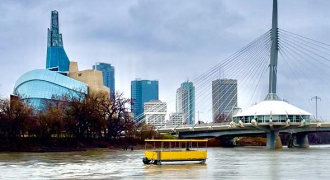 The Canadian Museum for Human Rights and a white bridge can be seen behind a yellow boat on a river.