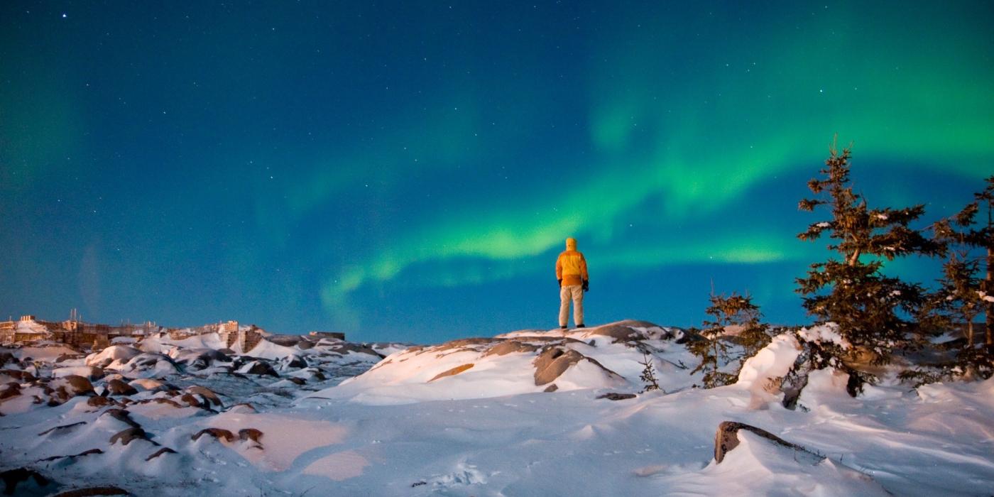A person standing on a snow covered ridge looking up at the green northern lights dancing across the sky.
