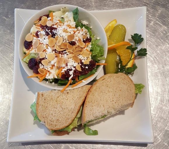 Frischer Salat mit Mandeln, ein geschnittenes Sandwich und in Scheiben geschnittene Gurken auf einem quadratischen Teller.