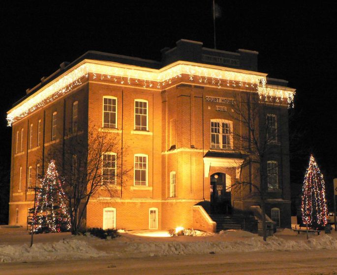 The Sam Waller Museum exterior lit up in winter with a spotlightt and holiday lights on the trees and roofline.